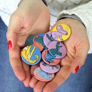 May include: A collection of colorful dinosaur and triceratops pinback buttons. The buttons are round and feature various dinosaurs in different colors, including blue, green, yellow, pink, and purple.