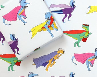 May include: White wrapping paper with a repeating pattern of cartoon dinosaurs dressed as superheroes. The dinosaurs are in blue, green, purple, and yellow, with pink, red, and purple capes. The paper is folded.