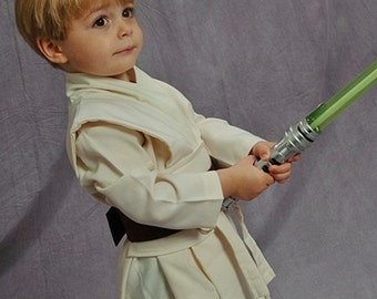 May include: A young child dressed in a white Jedi robe holds a green lightsaber. The child is looking to the right of the frame.
