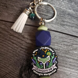 May include: A blue and green charm with the text "When life gets hard conjure up midnight margaritas" and a witch wearing a hat holding a margarita glass. The charm is attached to a silver key ring with a white tassel.