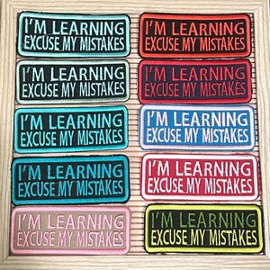 May include: A collection of rectangular embroidered patches in various colors, each bearing the text "I'M LEARNING EXCUSE MY MISTAKES" in a bold, contrasting font. The patches are arranged in a grid, showcasing a variety of color combinations.