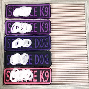 May include: Rectangular patches with embroidered text. The patches are in various colors, including purple, black, and pink, with white text. The text on the patches reads "SERVICE K9" and "SERVICE DOG". The patches are displayed in a wooden frame.