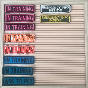 May include: A collection of embroidered patches with text, including "IN TRAINING" in various colors like pink, orange, and blue. Also, patches with "EMERGENCY INFO INSIDE" and "ASK TO PET" are visible. The patches are arranged on a light brown, ridged background.