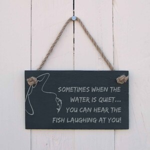 Könnte beinhalten: Ein schwarzes Schiefer-Schild mit einem weißen Seilhalter. Das Schild hat eine weiße Silhouette einer Angelrute und eines Fisches mit dem Text "SOMETIMES WHEN THE WATER IS QUIET... YOU CAN HEAR THE FISH LAUGHING AT YOU!"