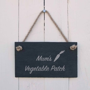 Op de afbeelding: Een zwart leisteen bord met een wit touw dat aan een spijker hangt. Het bord heeft de tekst "Mum's Vegetable Patch" met een kleine afbeelding van een wortel.