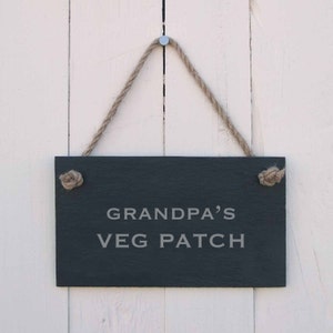 May include: A black slate sign with white text that reads "Grandpa's Veg Patch" hanging from a rope on a white wooden background.
