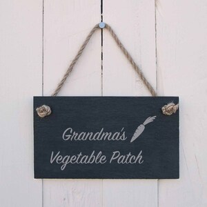 May include: A rectangular black slate sign with the text "Grandma's Vegetable Patch" and a carrot illustration. The sign is suspended by a natural rope and is attached to a white wooden background.