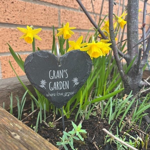 May include: A black heart-shaped slate garden sign with the text "Gran's Garden where love grows". The sign is placed in a planter with yellow daffodils and green plants.