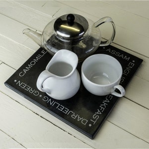 May include: A black slate serving tray with the words "Darjeeling", "Breakfast", "Assam", "English", "Green", and "Chamomile" printed on it. A glass teapot with a stainless steel lid and strainer is filled with tea. A white creamer and a white teacup are also on the tray.