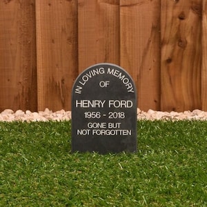 May include: A dark grey memorial stone with a rounded top, inscribed with "IN LOVING MEMORY OF HENRY FORD 1956-2018 GONE BUT NOT FORGOTTEN". The stone is set in green grass with a wooden fence in the background.
