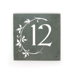 May include: A square slate house number sign with a white floral design and the number 12 in white.