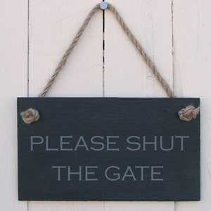 Op de afbeelding: Een zwart leisteen bord met een touw dat aan een spijker hangt. Het bord heeft de tekst "PLEASE SHUT THE GATE" in witte letters.