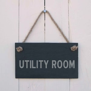 Può includere: Un cartello in ardesia nera con testo bianco che recita "UTILITY ROOM" appeso a una corda su una parete di legno bianco.