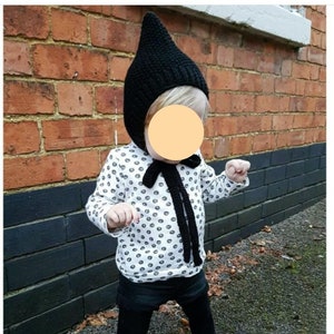 May include: A child wearing a black knitted pointed hat with a black and white polka dot shirt and black leggings. The child is standing in front of a brick wall.
