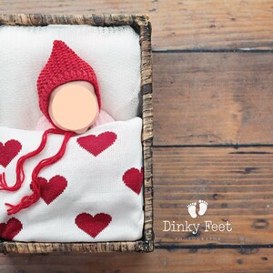 May include: A newborn baby wearing a red knitted hat with a long tassel, lying in a wicker basket lined with a white blanket with red hearts. The baby is sleeping peacefully.