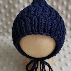May include: A navy blue knitted baby bonnet with a pointed top and a long tie. The bonnet is on a wooden mannequin head.