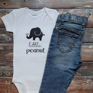 May include: White baby onesie with a black elephant graphic and the text "little peanut". A pair of blue denim jeans is next to the onesie.