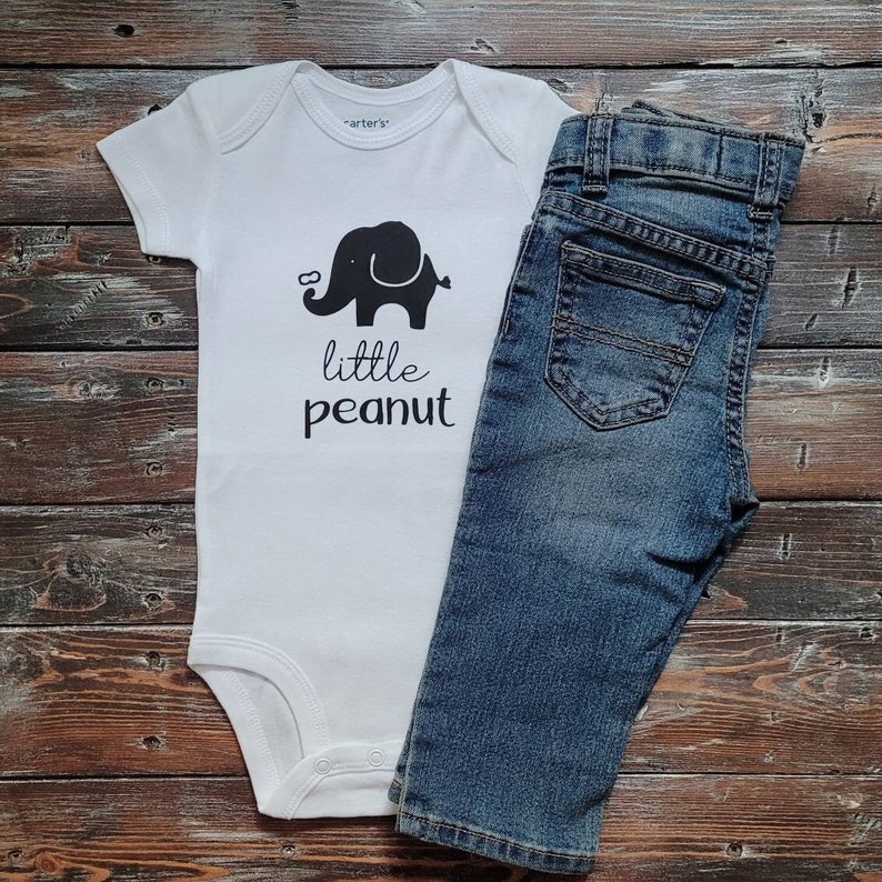 May include: White baby onesie with a black elephant graphic and the text "little peanut". A pair of blue denim jeans is next to the onesie.