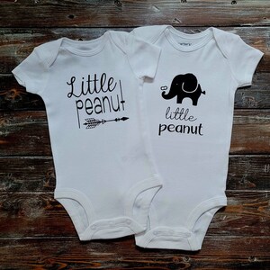 May include: Two white baby onesies with black text and graphics. One onesie says "Little Peanut" with an arrow pointing to the right. The other onesie says "Little Peanut" with a black silhouette of an elephant.