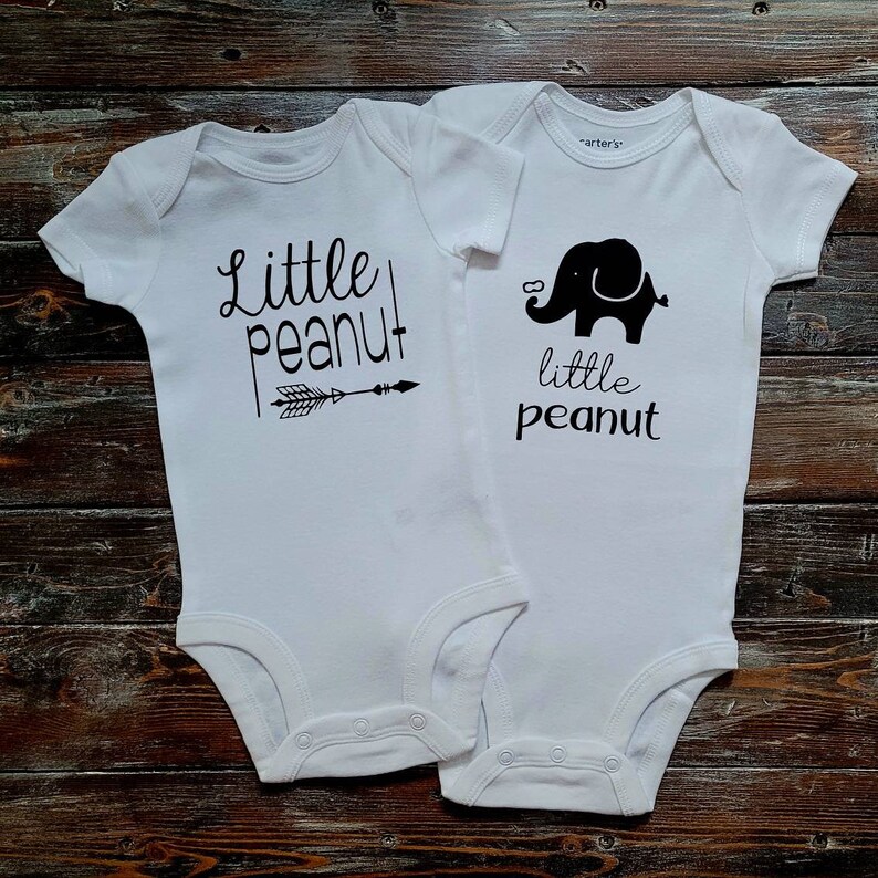 May include: Two white baby onesies with black text and graphics. One onesie says "Little Peanut" with an arrow pointing to the right. The other onesie says "Little Peanut" with a black silhouette of an elephant.