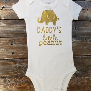 May include: White baby onesie with a gold elephant and the text "Daddy's little peanut".