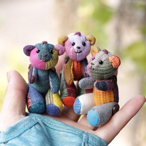 May include: Three small patchwork teddy bears in a hand. The bears are made of different colored fabrics, including pink, purple, blue, green, yellow, and orange. The bears have black eyes and black noses.