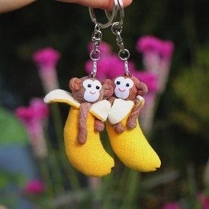 May include: Two keyrings with monkey plushies embracing yellow banana plushies. The monkeys have brown fur, white faces, and are attached to silver keyrings. The background is a blurred garden with pink flowers.
