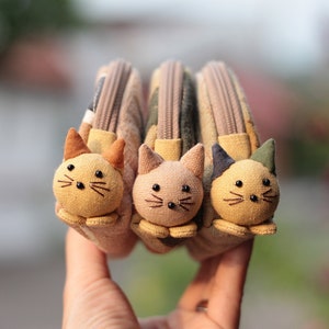 Può includere: Tre piccole borse di stoffa con facce di gatto. Le borse sono beige, verde e marrone con chiusure a zip.