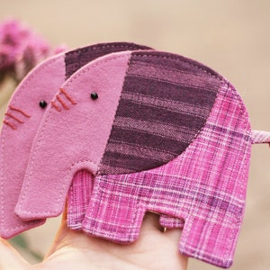 Pode incluir: Dois brinquedos de elefante de tecido rosa e roxo com detalhes costurados. Os elefantes são feitos de tecidos com padrões diferentes e têm uma cauda amarrada.