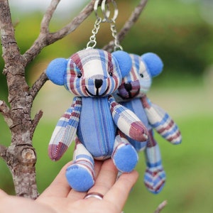 Könnte beinhalten: Zwei kleine Schlüsselanhänger in Form von Teddybären aus blau-weißem kariertem Stoff, die an einer silbernen Kette hängen. Die Bären halten sich an den Händen und haben blaue Filzohren und -nasen.