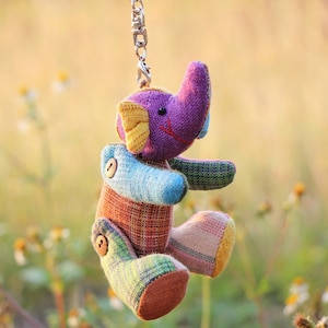 May include: A colorful patchwork elephant keychain with a purple head, yellow ears, and blue body. The keychain is hanging from a silver chain in a field of green grass.