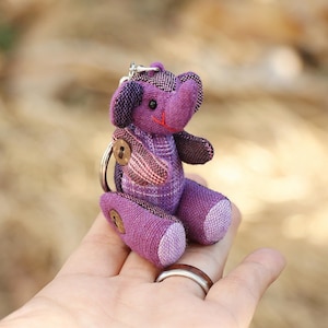 May include: Purple fabric elephant keychain with a button detail. The elephant is sitting on a hand.