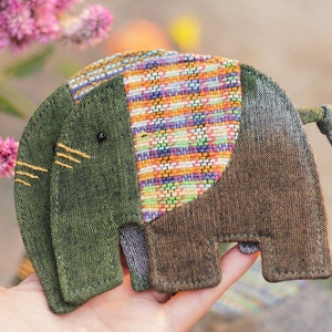 Puede incluir: Un pequeño elefante de peluche hecho a mano con tela de patchwork. El elefante es verde, marrón y multicolor con un ojo negro y costuras amarillas.