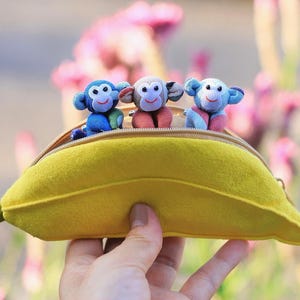 May include: A yellow banana-shaped zippered pouch featuring three small plush monkey toys peeking out from the top opening. The pouch has a green stem-like pull tab on one end and a soft, felt-like texture, with the monkeys in blue and brown.