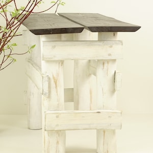 May include: A rustic wooden bench with two dark brown wooden planks as the seat and white painted legs. The legs are made of thick wooden beams with a distressed white paint finish.