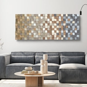 Puede incluir: Una gran pieza de arte mural rectangular de madera compuesta por numerosos bloques cuadrados pequeños en tonos beige, gris y blanco. Los bloques crean un efecto texturizado y degradado. La obra de arte cuelga sobre un sofá seccional gris.