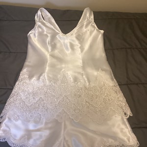 May include: White satin sleepwear with lace trim. The top has a V-neck and the shorts have a scalloped edge.