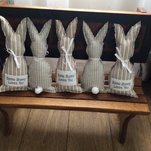 May include: Five brown and white striped bunny-shaped fabric pillows with white ribbon bows and the text "Some Bunny Loves You" on each pillow. The pillows are sitting on a wooden bench.
