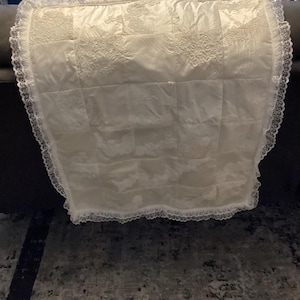 May include: A white satin baby quilt with a lace trim. The quilt is made of squares with embroidered designs.