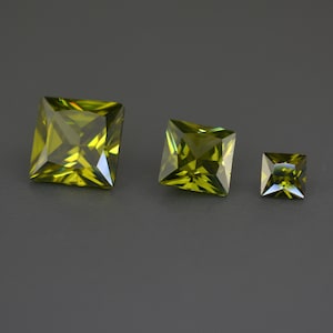May include: Three green square-shaped gemstones of different sizes. The gemstones are faceted and have a slight yellow hue.