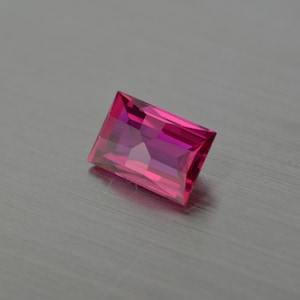 Op de afbeelding: Een rechthoekige, gefacetteerde edelsteen met een roze tot paarse tint. De edelsteen is geplaatst tegen een zilveren achtergrond.