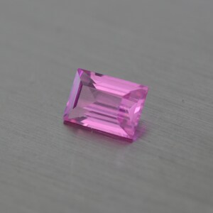 May include: A faceted, rectangular pink gemstone with a step cut. The gem is a vibrant shade of pink and appears to be transparent. The gemstone is set against a neutral, gray background.