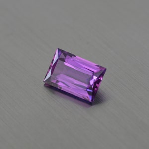 Puede incluir: Una piedra preciosa rectangular, facetada, de color púrpura con un tono púrpura claro.