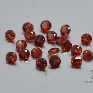 May include: A collection of round, red gemstones. The gemstones are scattered on a white surface.