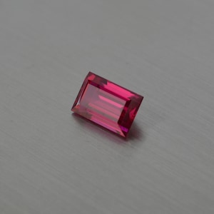 Peut inclure: Une pierre précieuse rectangulaire facetée de couleur rose. La pierre précieuse est placée sur un fond gris.