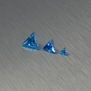 Op de afbeelding: Drie blauwe, driehoekige edelstenen van verschillende grootte op een geborsteld zilveren oppervlak.