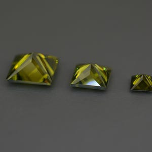 May include: Three green square-shaped gemstones of different sizes. The gemstones are faceted and have a pyramid shape.