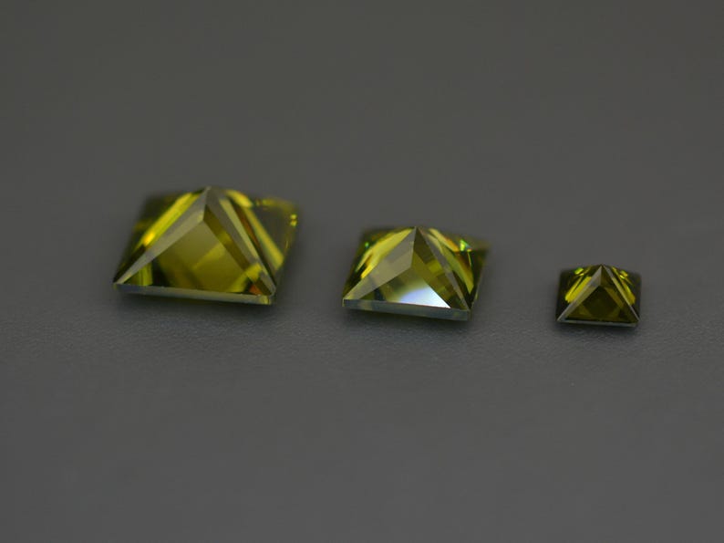 May include: Three green square-shaped gemstones of different sizes. The gemstones are faceted and have a pyramid shape.