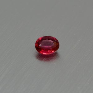 Puede incluir: Una gema ovalada de color rojo vibrante con una superficie lisa y pulida.