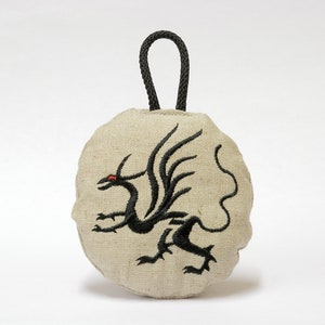 May include: A round, beige linen bag with a black drawstring closure. The bag features a black embroidered dragon with red eyes.